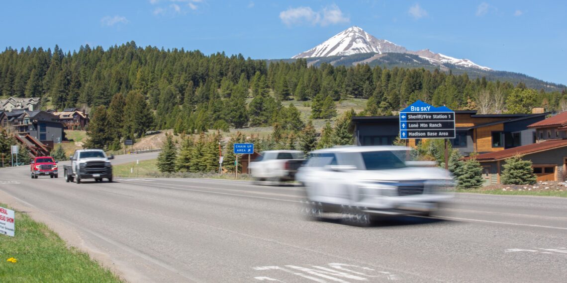 Two cars drive on a multi-lane road with a forested hillside and a snow‑capped mountain in the distance.