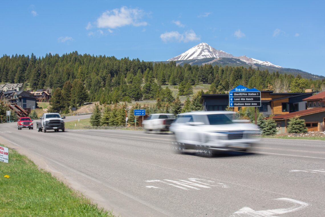 Two cars drive on a multi-lane road with a forested hillside and a snow‑capped mountain in the distance.