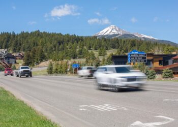 Two cars drive on a multi-lane road with a forested hillside and a snow‑capped mountain in the distance.