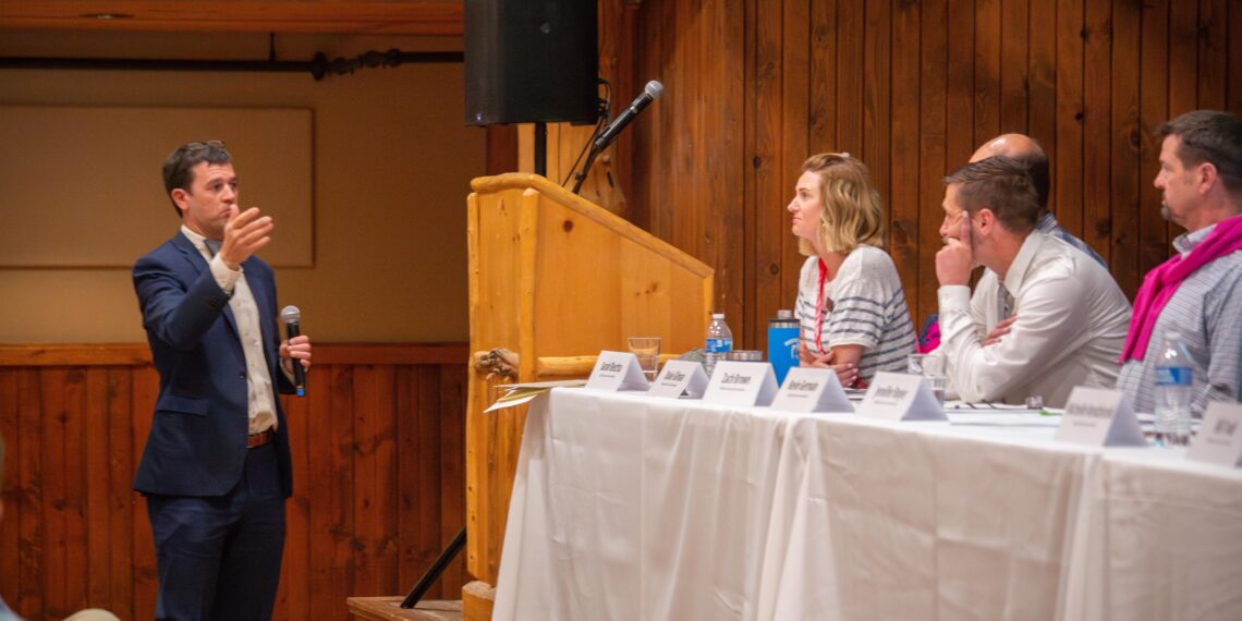 Man in a blue suit speaks into a handheld microphone beside a wooden podium at a panel discussion by the stage.