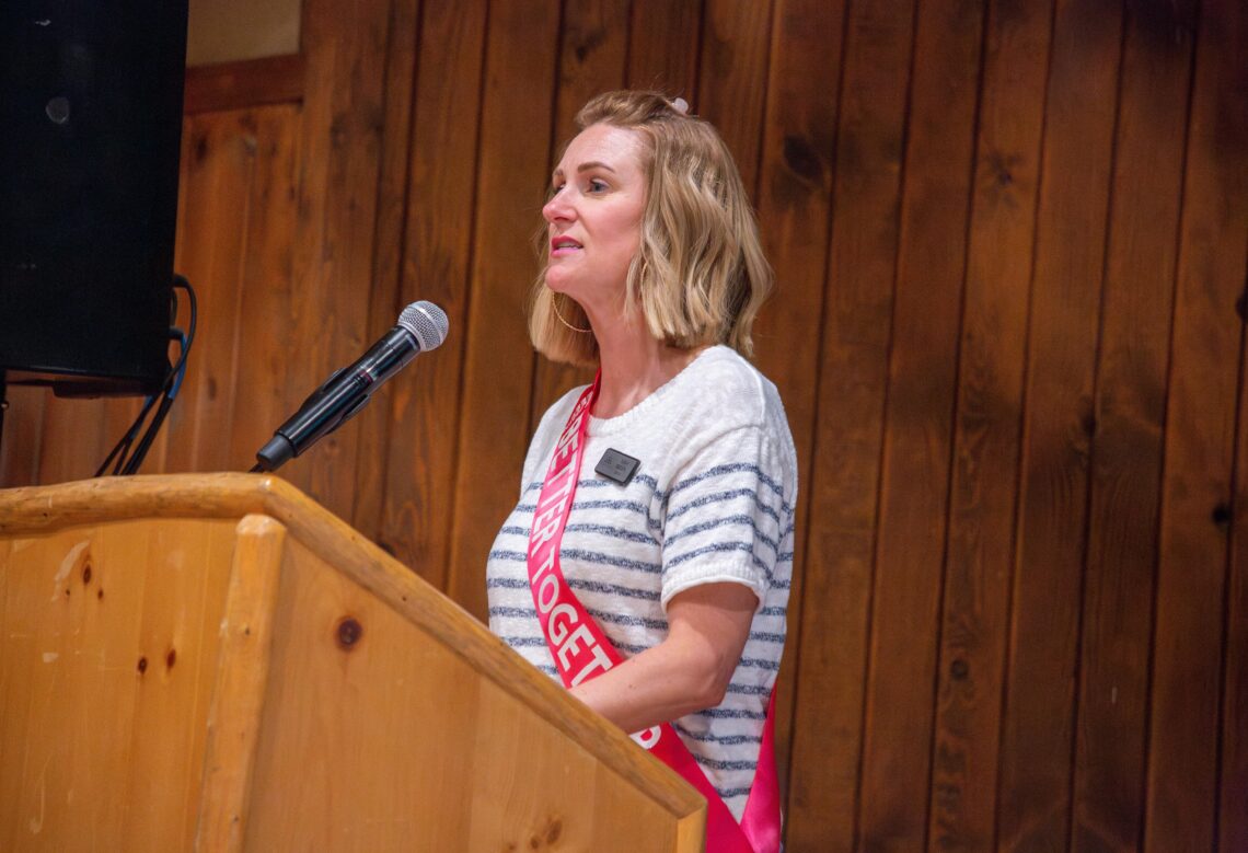 Woman with shoulder-length blonde hair speaks into a microphone at a wooden podium, wearing a striped shirt and pink sash.