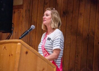 Woman with shoulder-length blonde hair speaks into a microphone at a wooden podium, wearing a striped shirt and pink sash.