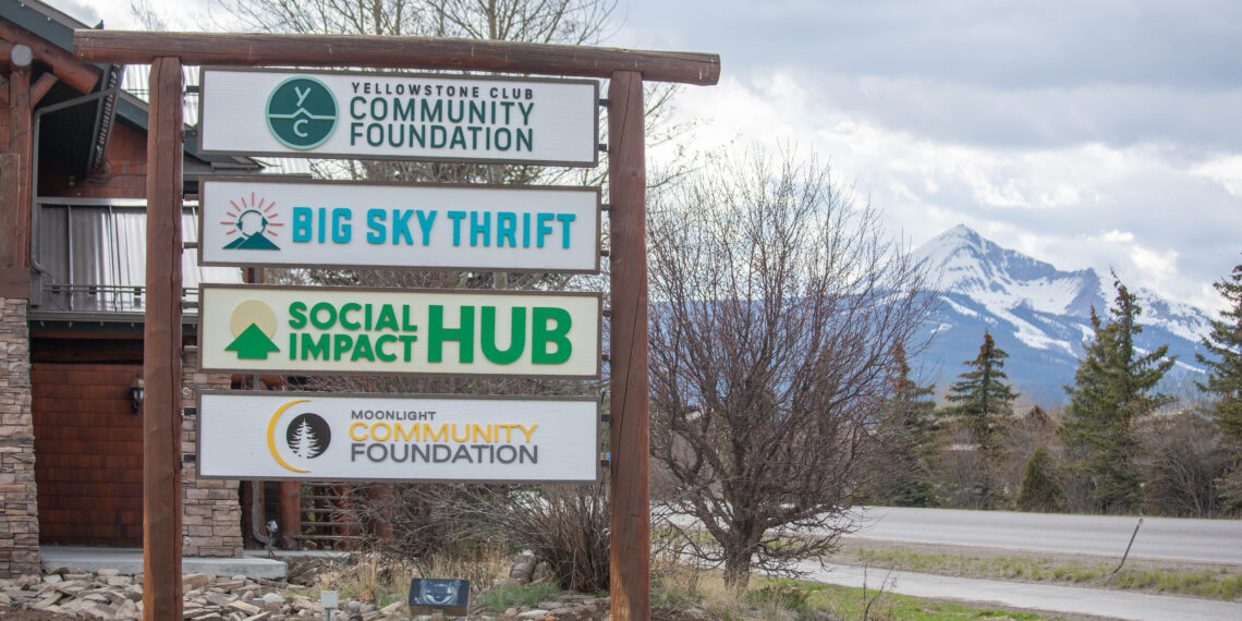 Signpost with multiple community organization signs against mountain backdrop (Big Sky Thrift, Community Foundation, Social Hub)