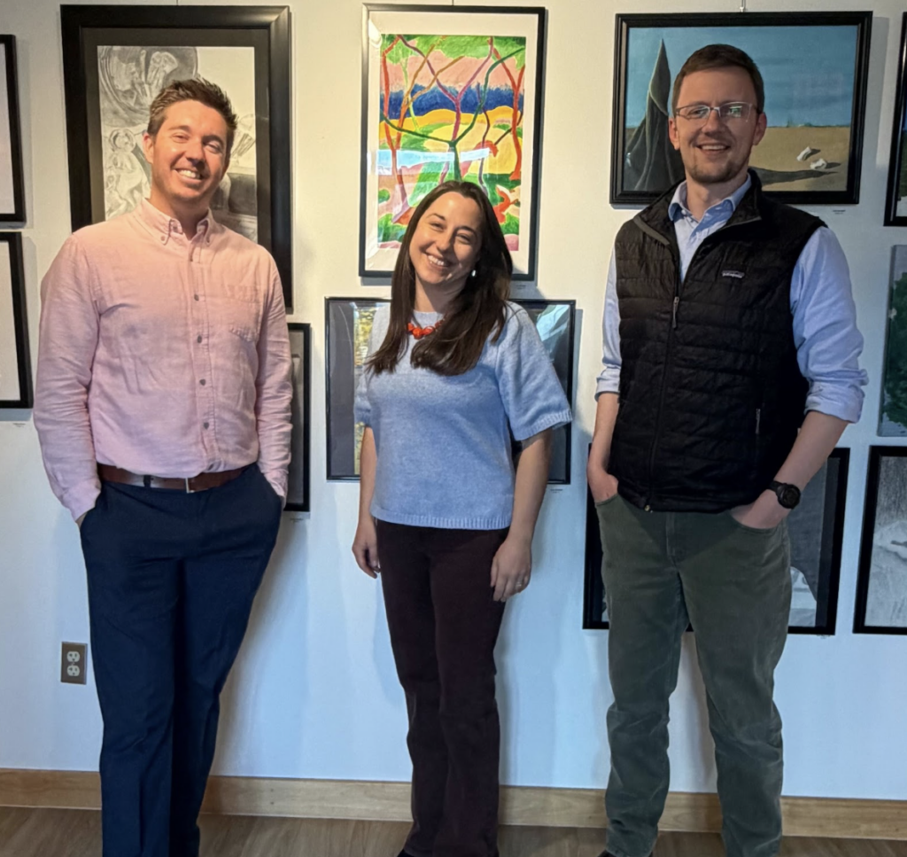 Three adults stand side by side in an art gallery, smiling at the camera, with framed artwork on the wall behind them.