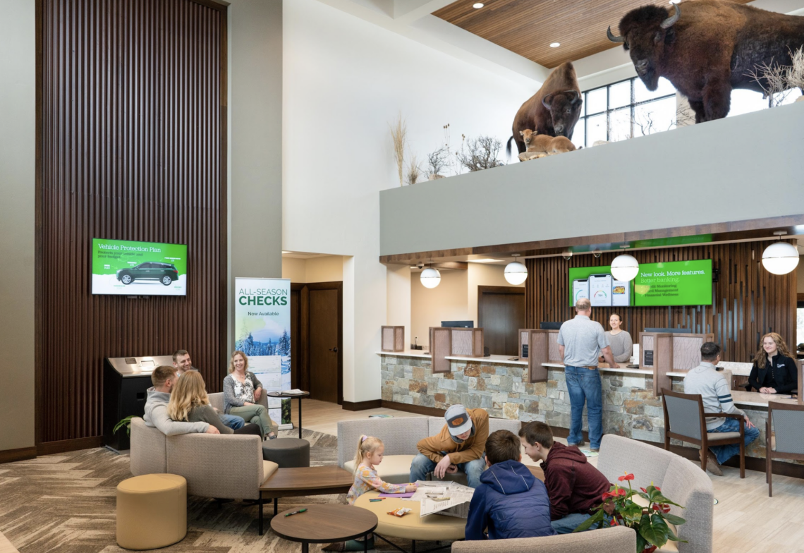 Modern bank lobby with seating areas and customers at service desks, plus buffalo statues visible on an upper level.