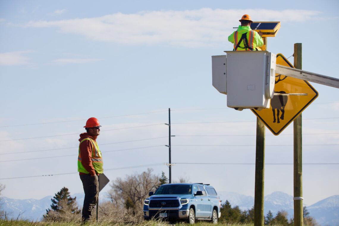 Worker in neon safety vest and orange hard hat stands beside a pickup truck by a road sign; another worker in a bucket truck inspects equipment at an elevated platform with a solar panel setup behind him.