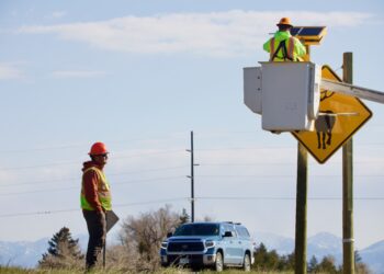 Worker in neon safety vest and orange hard hat stands beside a pickup truck by a road sign; another worker in a bucket truck inspects equipment at an elevated platform with a solar panel setup behind him.