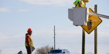 Worker in neon safety vest and orange hard hat stands beside a pickup truck by a road sign; another worker in a bucket truck inspects equipment at an elevated platform with a solar panel setup behind him.