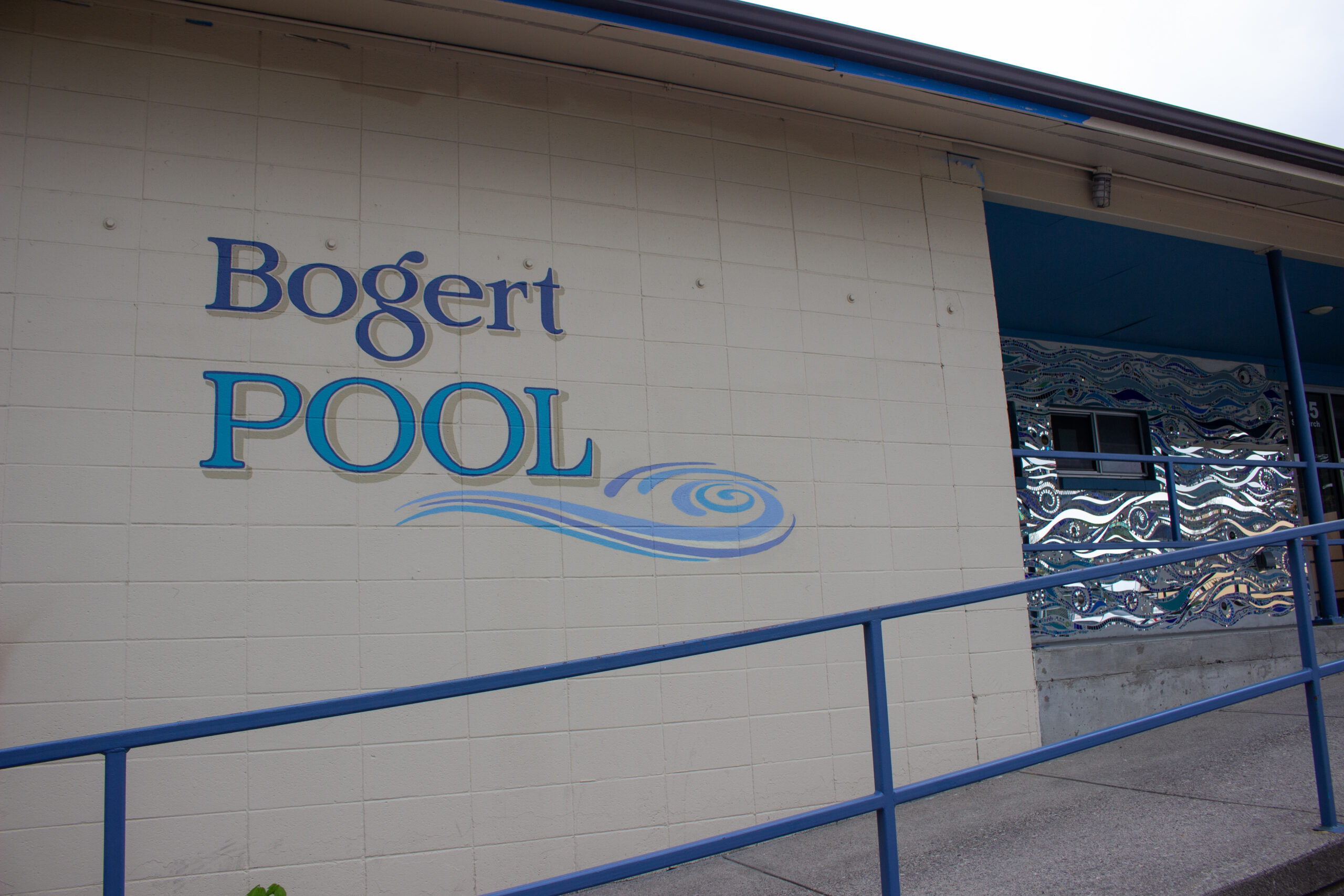 Exterior wall of Bogert Pool with blue sign lettering and decorative wave mural; blue railing in foreground.