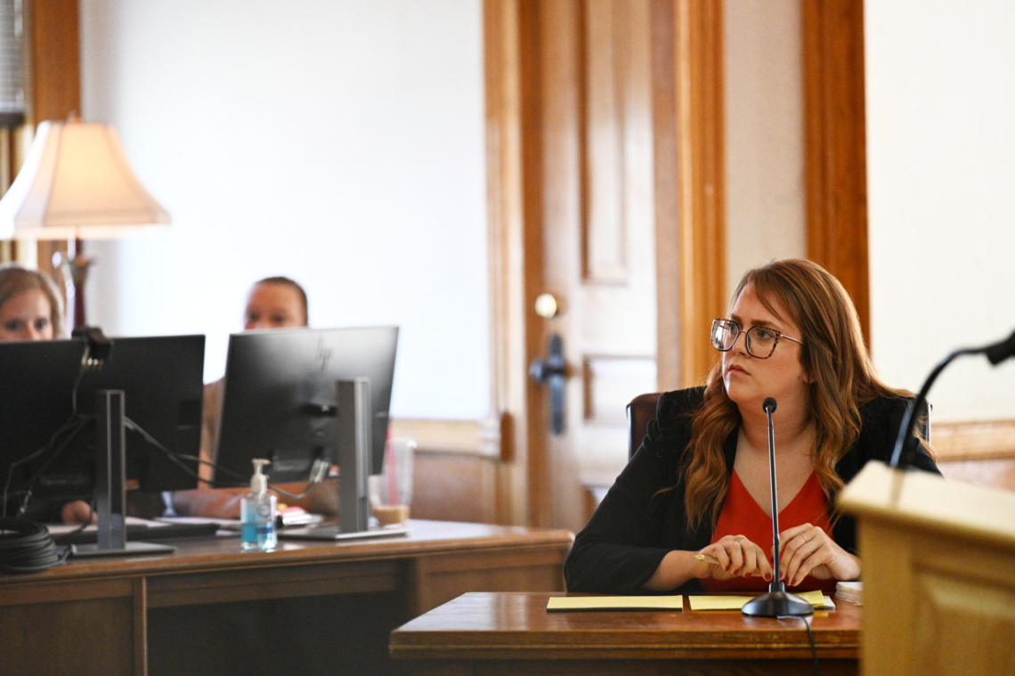 A woman with glasses speaking into a microphone at a wooden conference table, colleagues with monitors in the background.