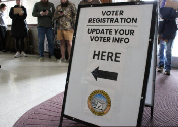 Bold voter registration sign in a public space reading 'Voter Registration Update Your Voter Info Here' with a left-pointing arrow and county seal visible below.