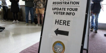 Bold voter registration sign in a public space reading 'Voter Registration Update Your Voter Info Here' with a left-pointing arrow and county seal visible below.