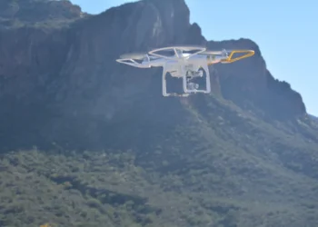 White quadcopter drone with protective guards hovering over a rugged mountain landscape under a clear blue sky