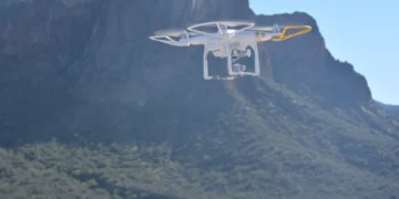 White quadcopter drone with protective guards hovering over a rugged mountain landscape under a clear blue sky