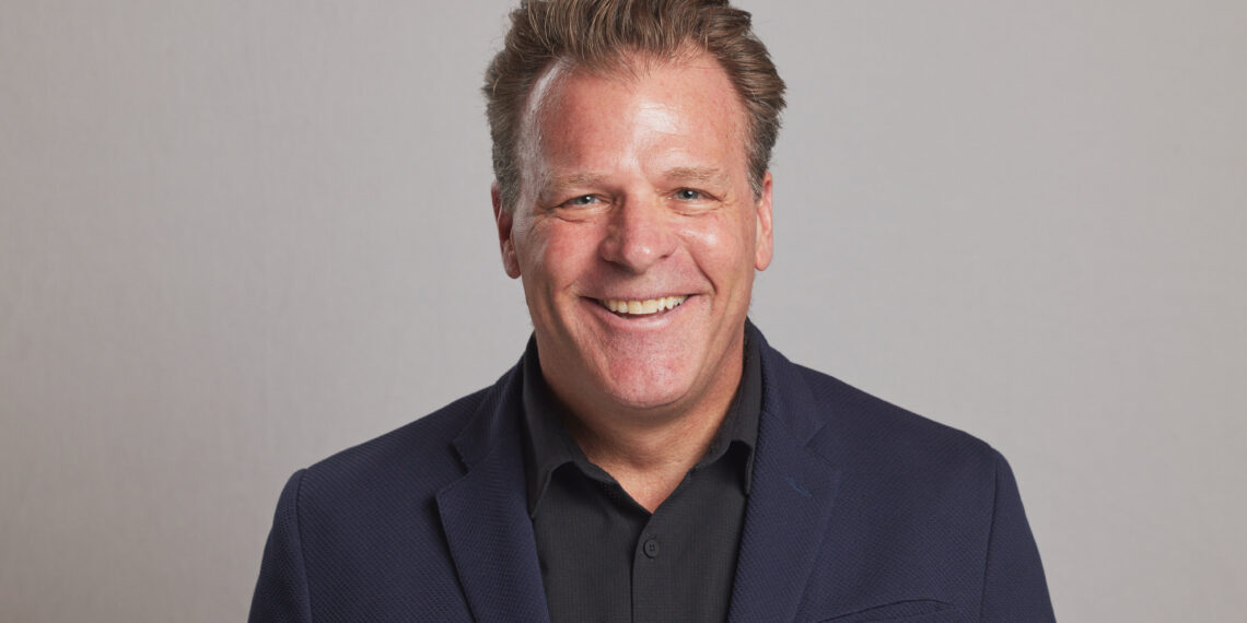 Smiling man in a dark blazer and black shirt against a neutral gray background (head and shoulders).
