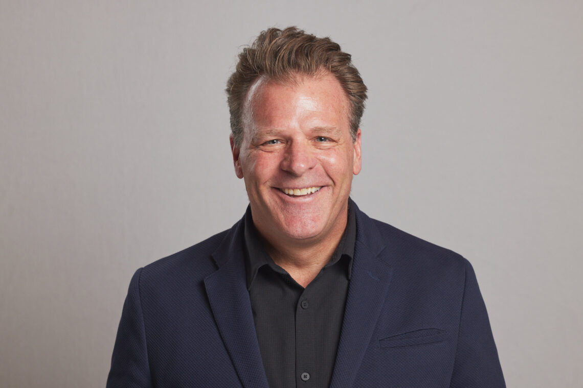 Smiling man in a dark blazer and black shirt against a neutral gray background (head and shoulders).