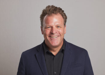 Smiling man in a dark blazer and black shirt against a neutral gray background (head and shoulders).