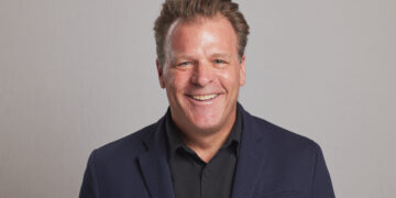 Smiling man in a dark blazer and black shirt against a neutral gray background (head and shoulders).