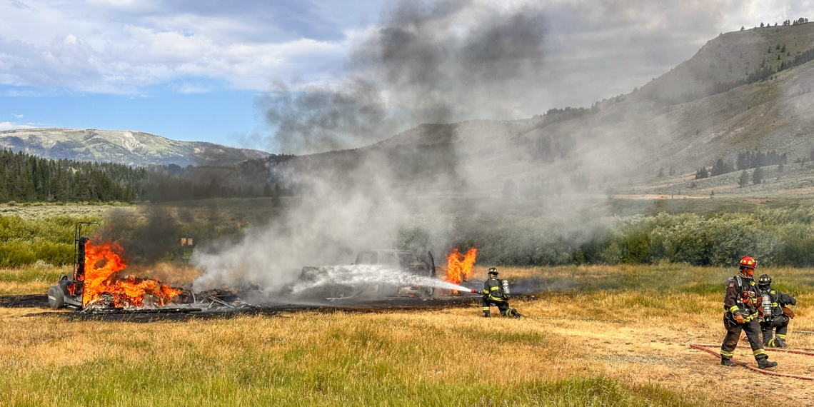 Firefighters spray water on a burning vehicle in a grassy field, with smoke rising and hills in the background.