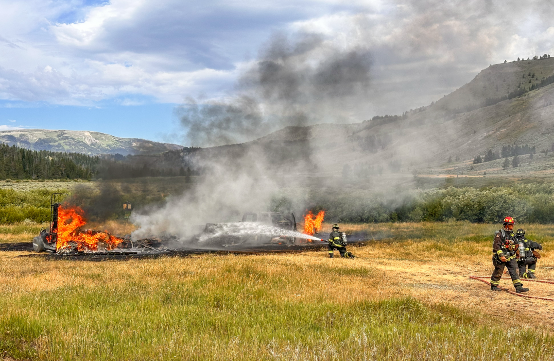 Firefighters spray water on a burning vehicle in a grassy field, with smoke rising and hills in the background.