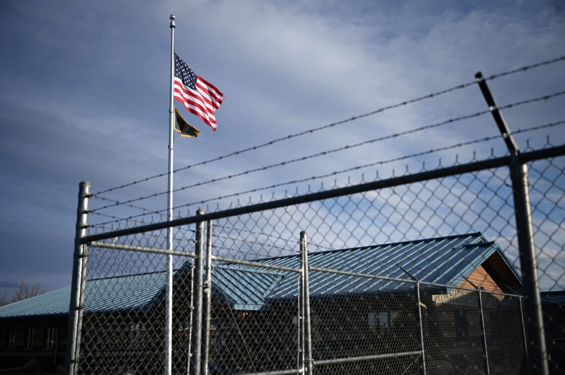 American flag and a second flag waving on a tall pole beside a barbed-wire fence and industrial buildings under a blue sky.