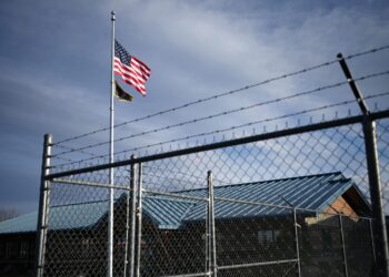 American flag and a second flag waving on a tall pole beside a barbed-wire fence and industrial buildings under a blue sky.