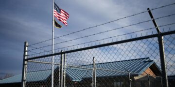 American flag and a second flag waving on a tall pole beside a barbed-wire fence and industrial buildings under a blue sky.