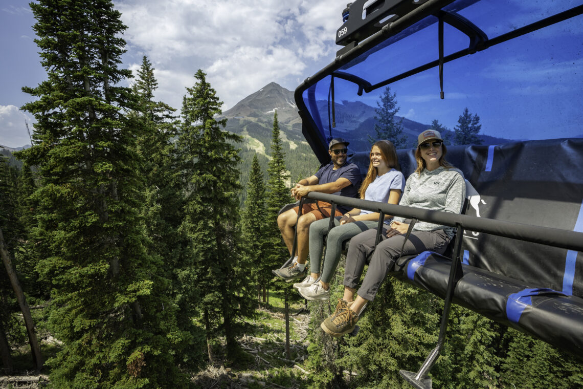 Three friends ride a blue chairlift above a forested valley with a distant mountain peak behind them