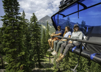 Three friends ride a blue chairlift above a forested valley with a distant mountain peak behind them