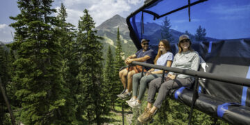 Three friends ride a blue chairlift above a forested valley with a distant mountain peak behind them