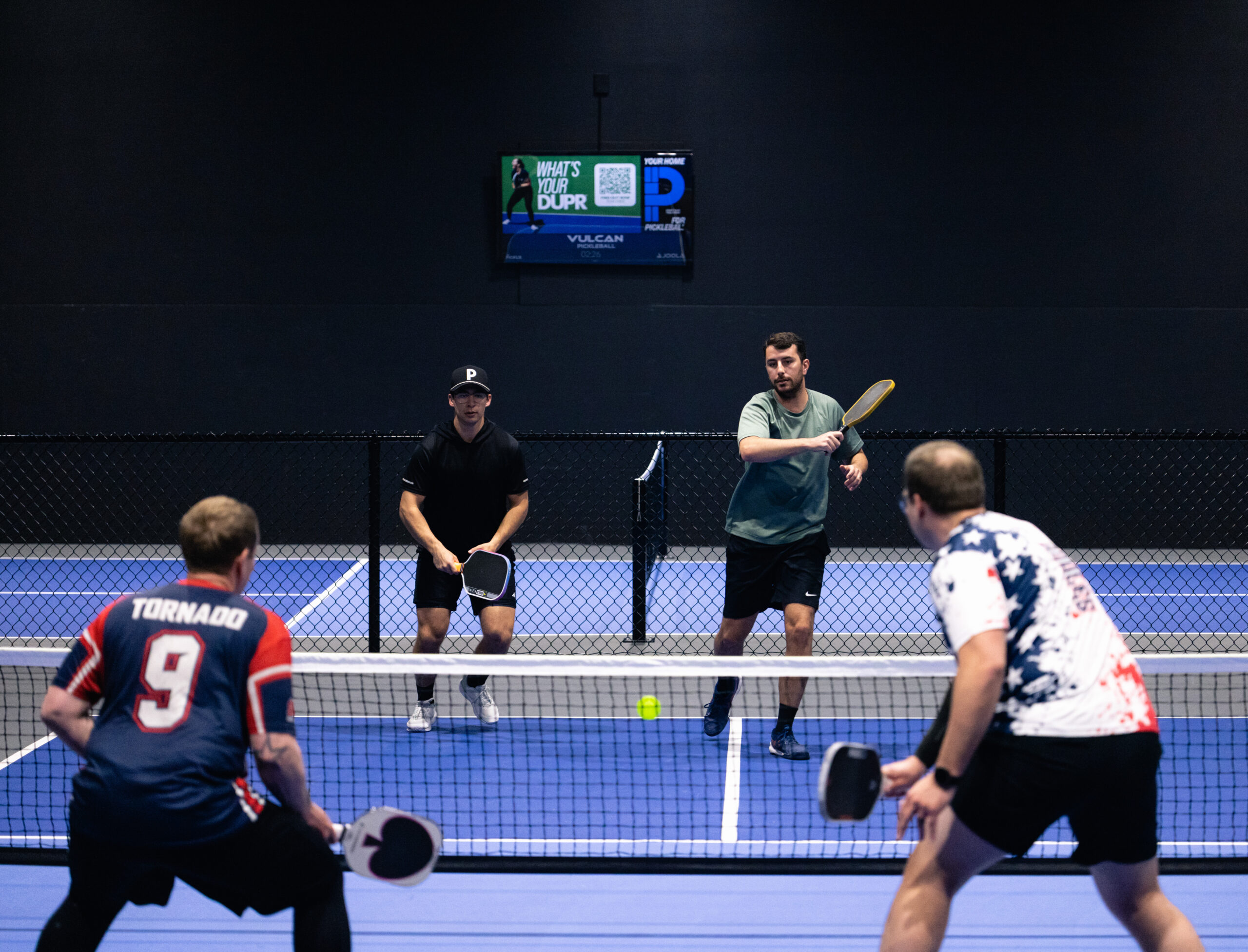Four players on a blue pickleball court behind a net, two facing each other with paddles as a ball flies between them indoors behind a dark wall.