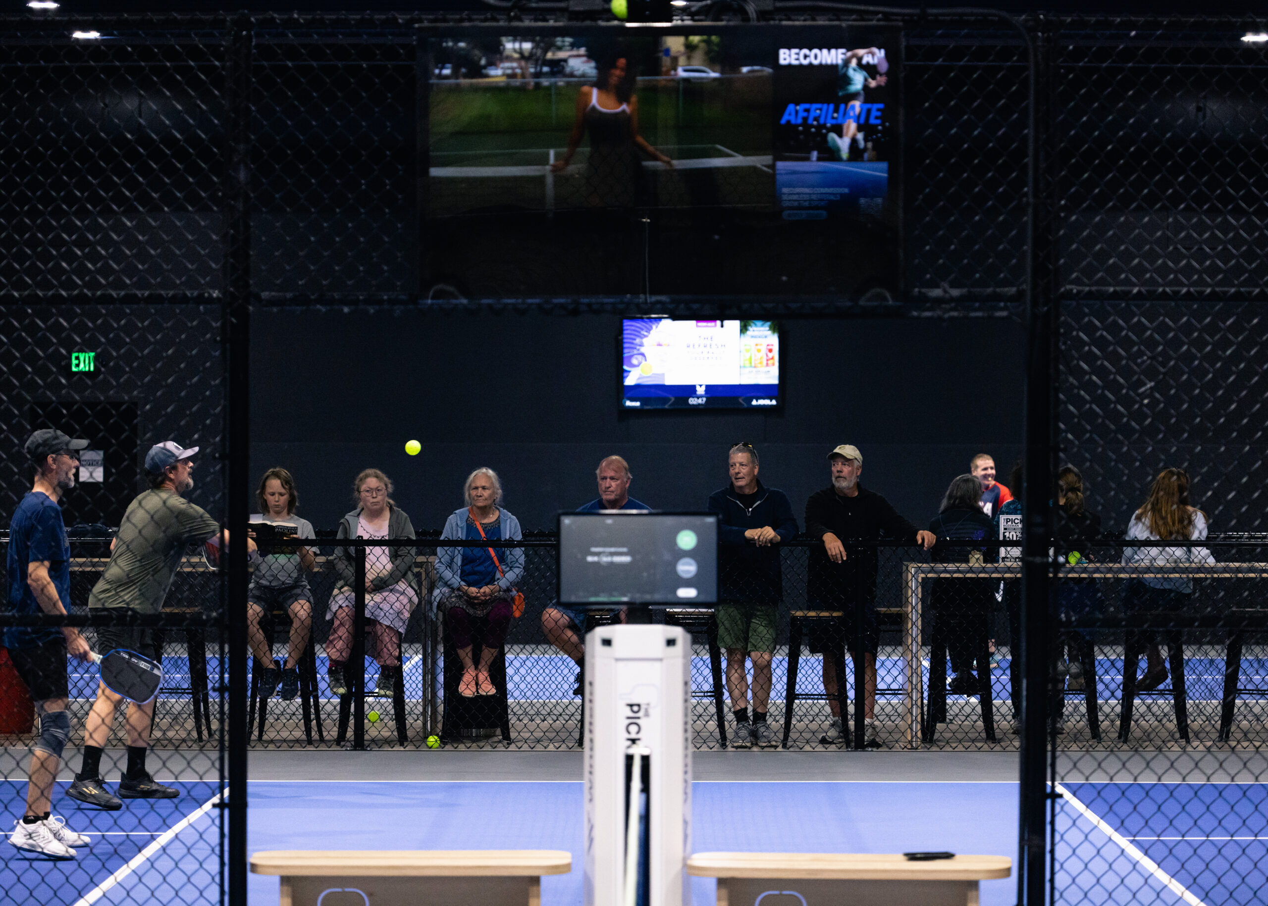 Group of spectators behind a chain-link fence watching a tennis match on a blue court.
