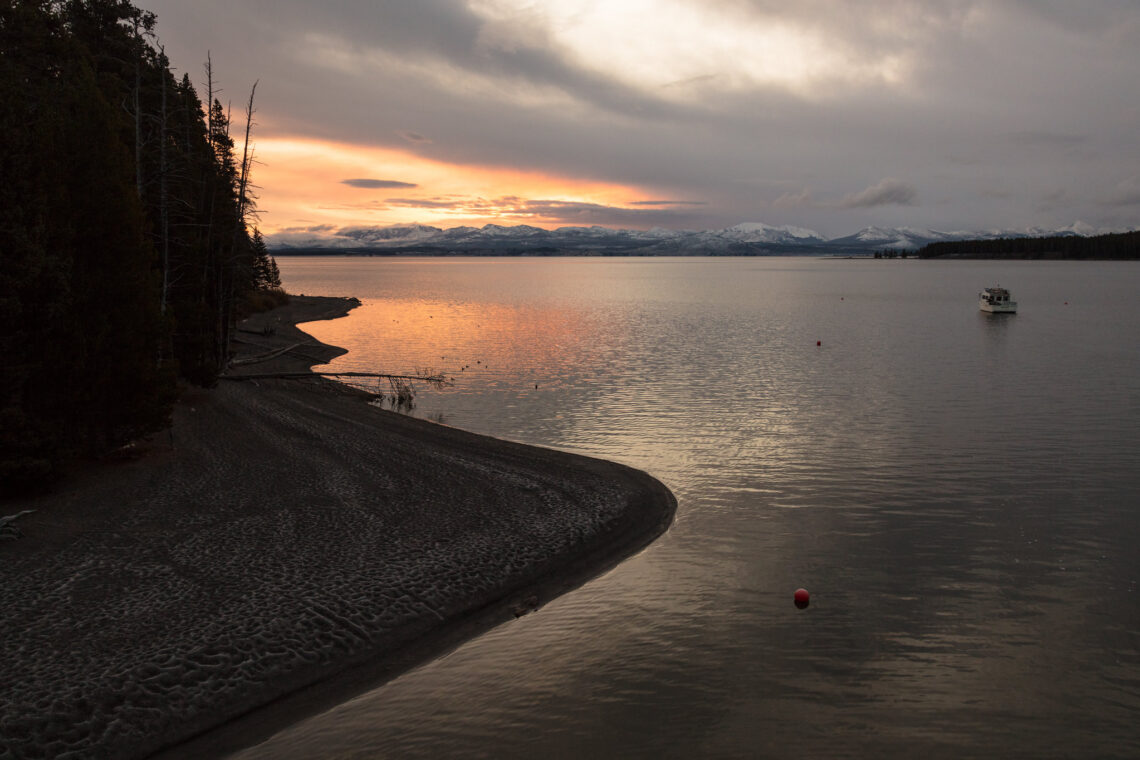 Sunset over a calm lake with snow-capped mountains in the distance; a small boat on the right and a rocky shoreline on the left.