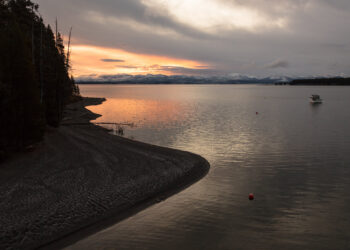 Sunset over a calm lake with snow-capped mountains in the distance; a small boat on the right and a rocky shoreline on the left.