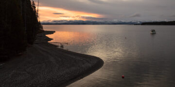 Sunset over a calm lake with snow-capped mountains in the distance; a small boat on the right and a rocky shoreline on the left.