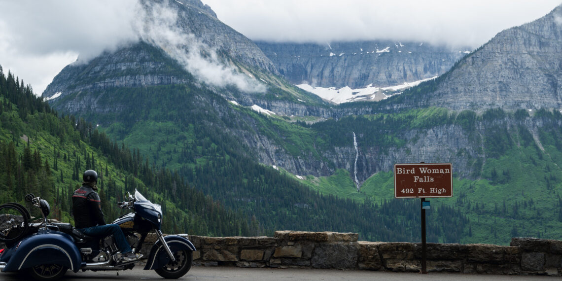 Motorcycle parked by a stone railing with a rider in a helmet, mountains in the background and a sign reading Bird Woman Falls 492 ft high, over a forested valley.