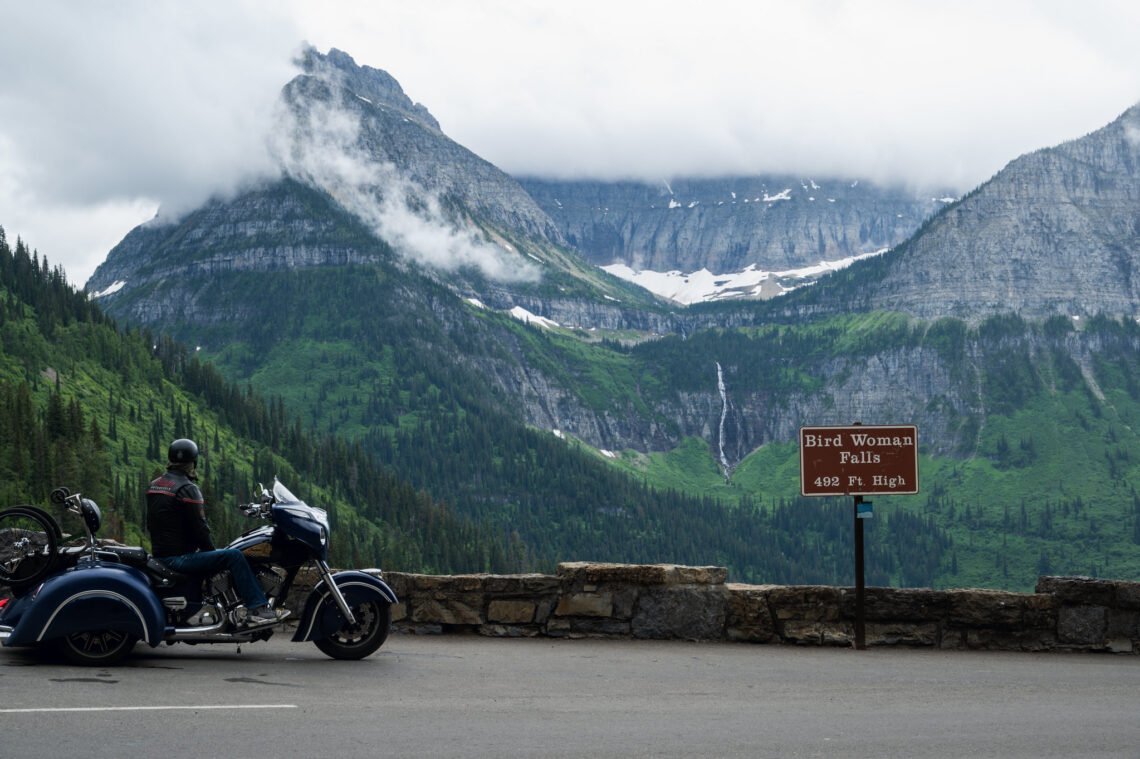 Motorcycle parked by a stone railing with a rider in a helmet, mountains in the background and a sign reading Bird Woman Falls 492 ft high, over a forested valley.