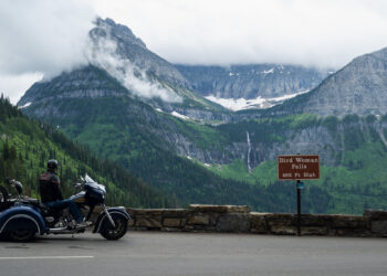 Motorcycle parked by a stone railing with a rider in a helmet, mountains in the background and a sign reading Bird Woman Falls 492 ft high, over a forested valley.
