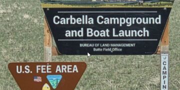 Sign for Carbella Campground and Boat Launch managed by the Bureau of Land Management (Butte Field Office) in a grassy field beneath a painted top panel image.
