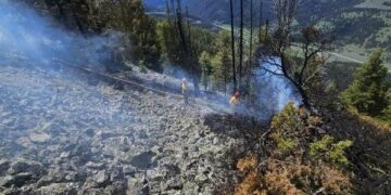 Rocky hillside with smoke wisps, two workers in yellow helmets, and distant green valley under a blue sky; wildfire on rocky slope.