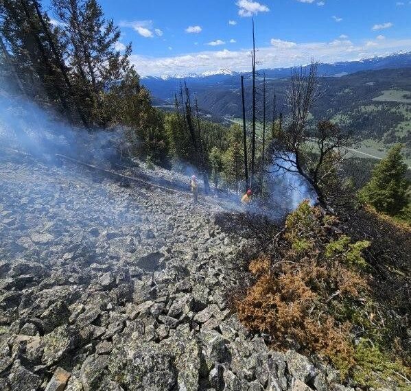 Rocky hillside with smoke wisps, two workers in yellow helmets, and distant green valley under a blue sky; wildfire on rocky slope.