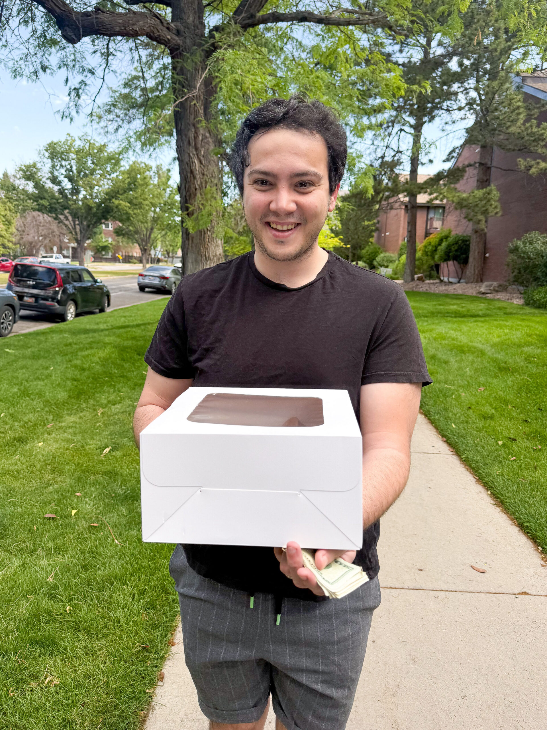 Smiling man in a black T-shirt holds a white box with chocolate cake visible through a window, standing on a sunny suburban sidewalk.