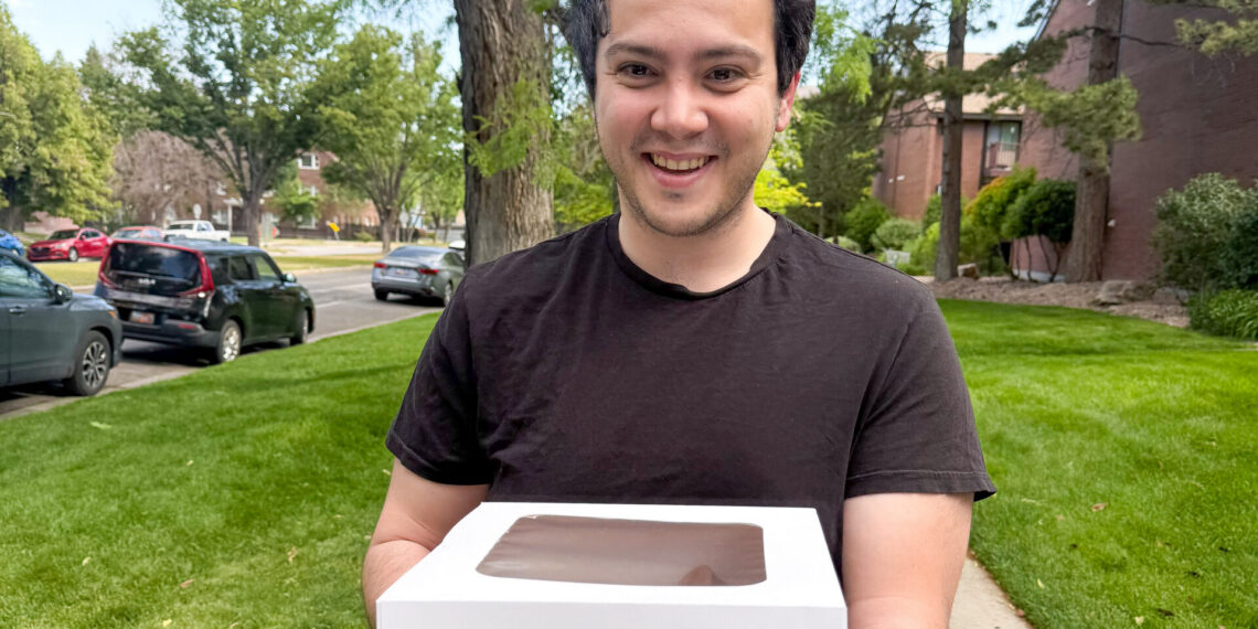 Smiling man in a black t-shirt holds a white cake box with a chocolate-topped window outdoors.