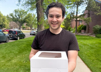 Smiling man in a black t-shirt holds a white cake box with a chocolate-topped window outdoors.