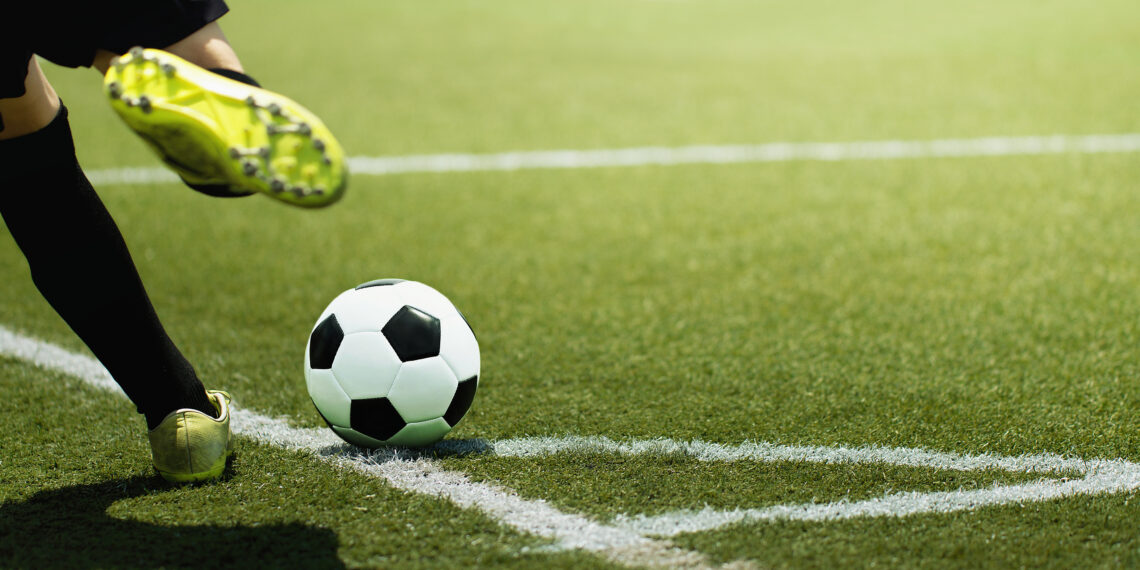 Soccer ball on a green turf near the sideline as a player strikes it with a yellow cleat