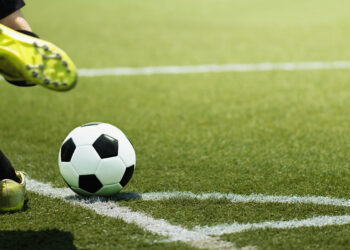 Soccer ball on a green turf near the sideline as a player strikes it with a yellow cleat