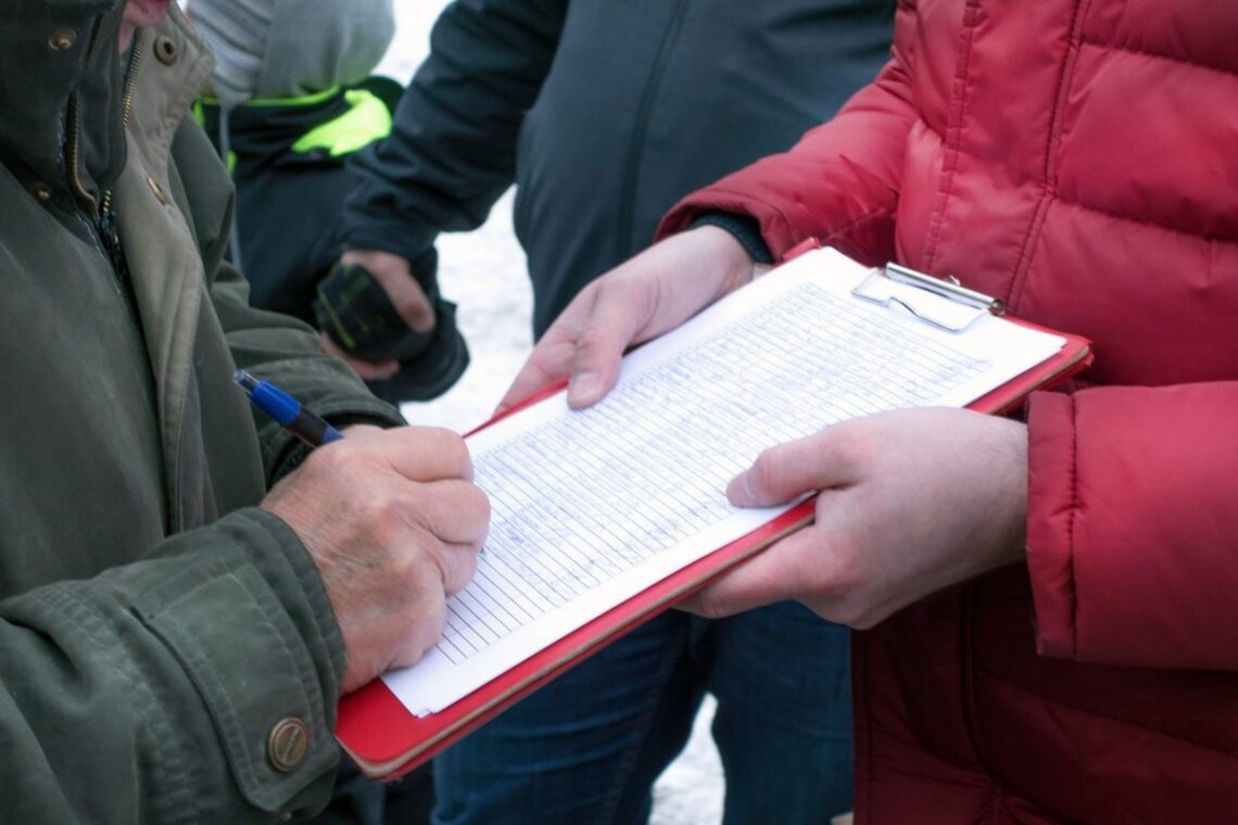 Person in a green jacket signs a clipboard held by someone in a red jacket outdoors.
