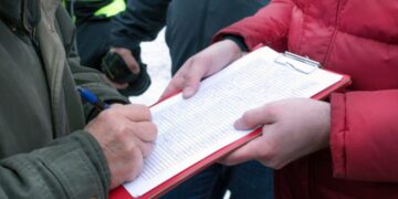 Person in a green jacket signs a clipboard held by someone in a red jacket outdoors.