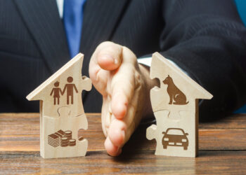 Hands pressing together two wooden house-shaped puzzle pieces, one with a family icon and coins, the other with a car icon, symbolizing family, housing, and financial planning.