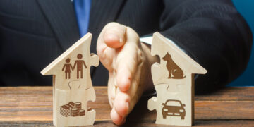 Hands pressing together two wooden house-shaped puzzle pieces, one with a family icon and coins, the other with a car icon, symbolizing family, housing, and financial planning.